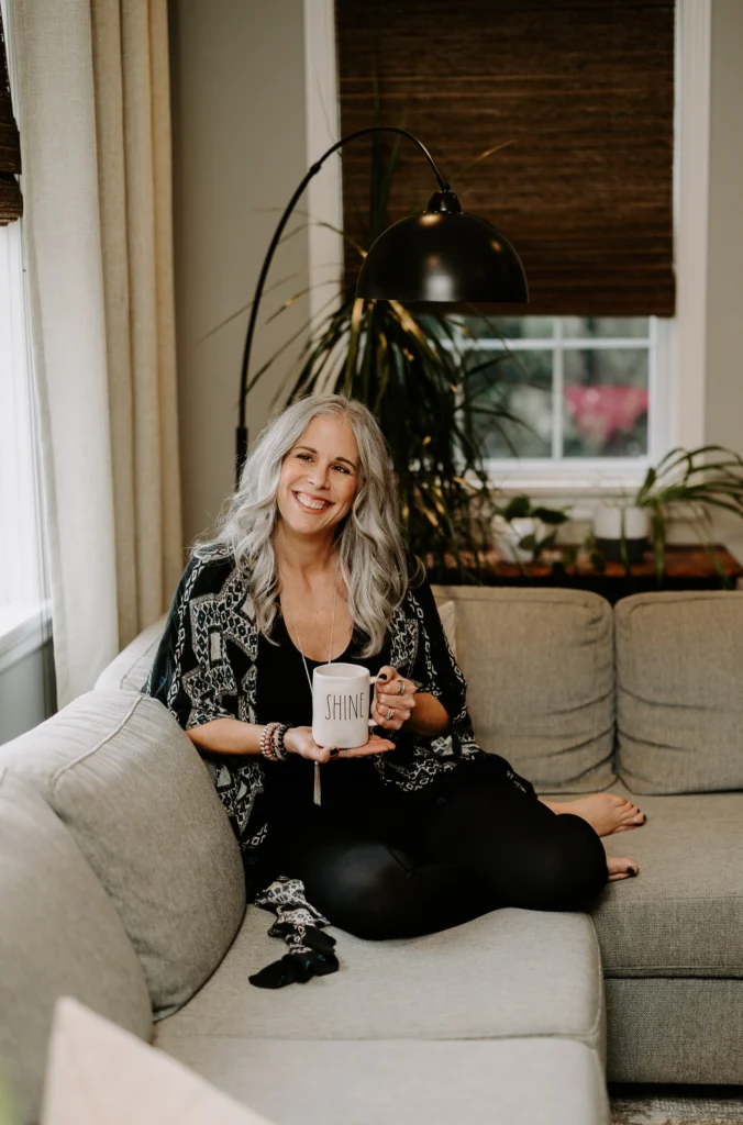 Dawne Sullivan, Holistic Nutritionist, Reiki Master, Cold Immersion Coach, Breathwork Facilitator sitting on couch with Shine mug smiling